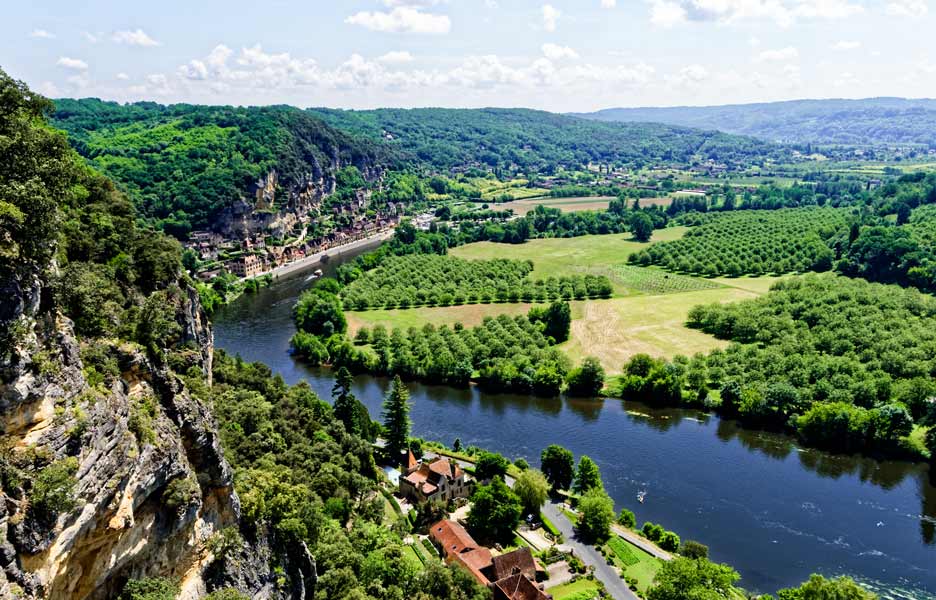 Vue panoramique aérienne d’une vallée verdoyante avec une rivière bordée de falaises rocheuses, un village au bord de l’eau et des champs et forêts s’étendant jusqu’aux collines.