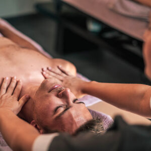 Masseur effectuant une séance de massage sur le dos d’un homme allongé, les mains posées sur le torse et le massage du visage en arrière-plan.