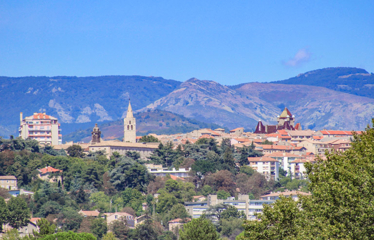 Vue panoramique d’une ville de montagne avec des églises et des maisons au pied de collines, entourée de végétation et de reliefs bleutés sous un ciel bleu.