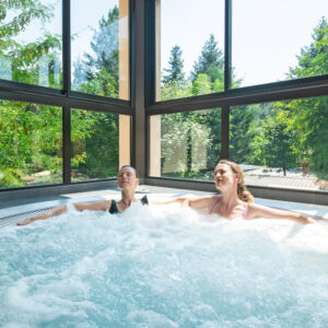 Deux femmes se détendent dans une piscine intérieure à remous, la tête émergeant de l’eau, fenêtres encadrant un paysage de verdure.