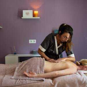 Thérapeute en uniforme de massage pratiquant un soin du dos sur une cliente allongée sur une table, dans une salle de soins aux murs mauves, avec bougies et étagères en arrière-plan.