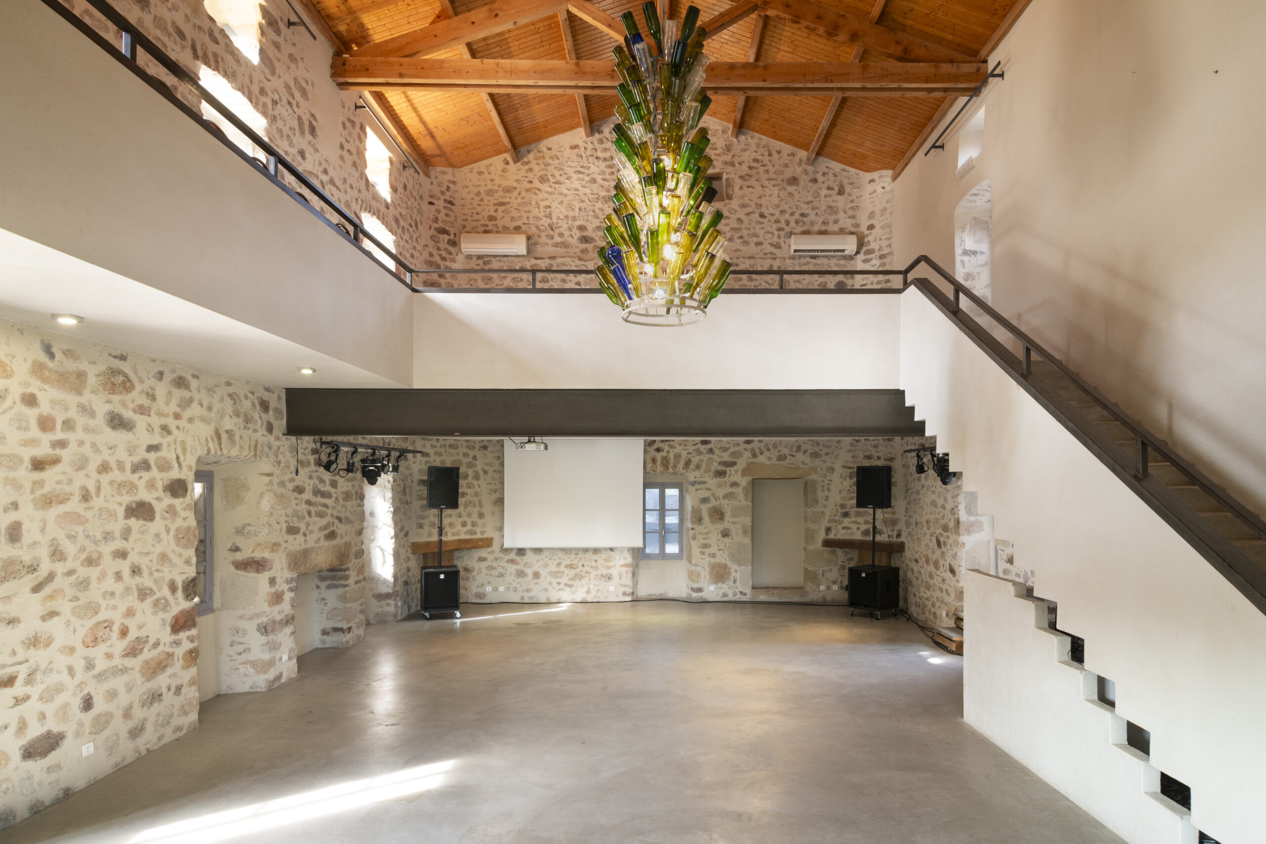 Grand hall intérieur avec murs en pierres apparentes, poutres en bois au plafond et lustre en verre coloré au centre, escaliers avec rampe sombre à droite, scène vide au fond avec écran de projection et deux enceintes sur pieds.
