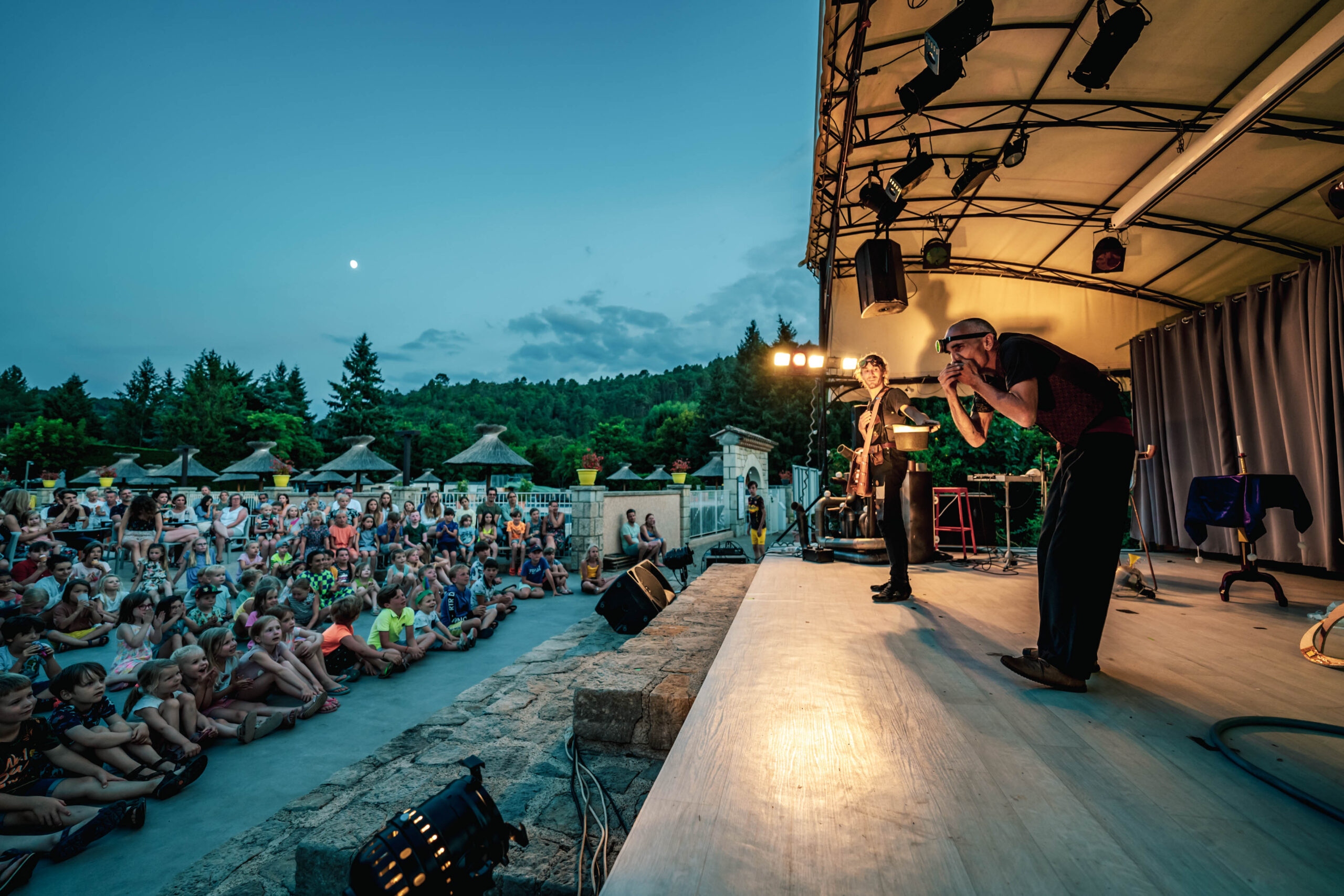 Scène en plein air au crépuscule : un musicien chante et joue devant un public d’enfants et de familles assis, sous un chapiteau avec éclairages et projecteurs.