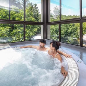 Couple souriant installé dans un bain à remous intérieur, avec bulles de mousse et vue sur la verdure à travers de grandes fenêtres.