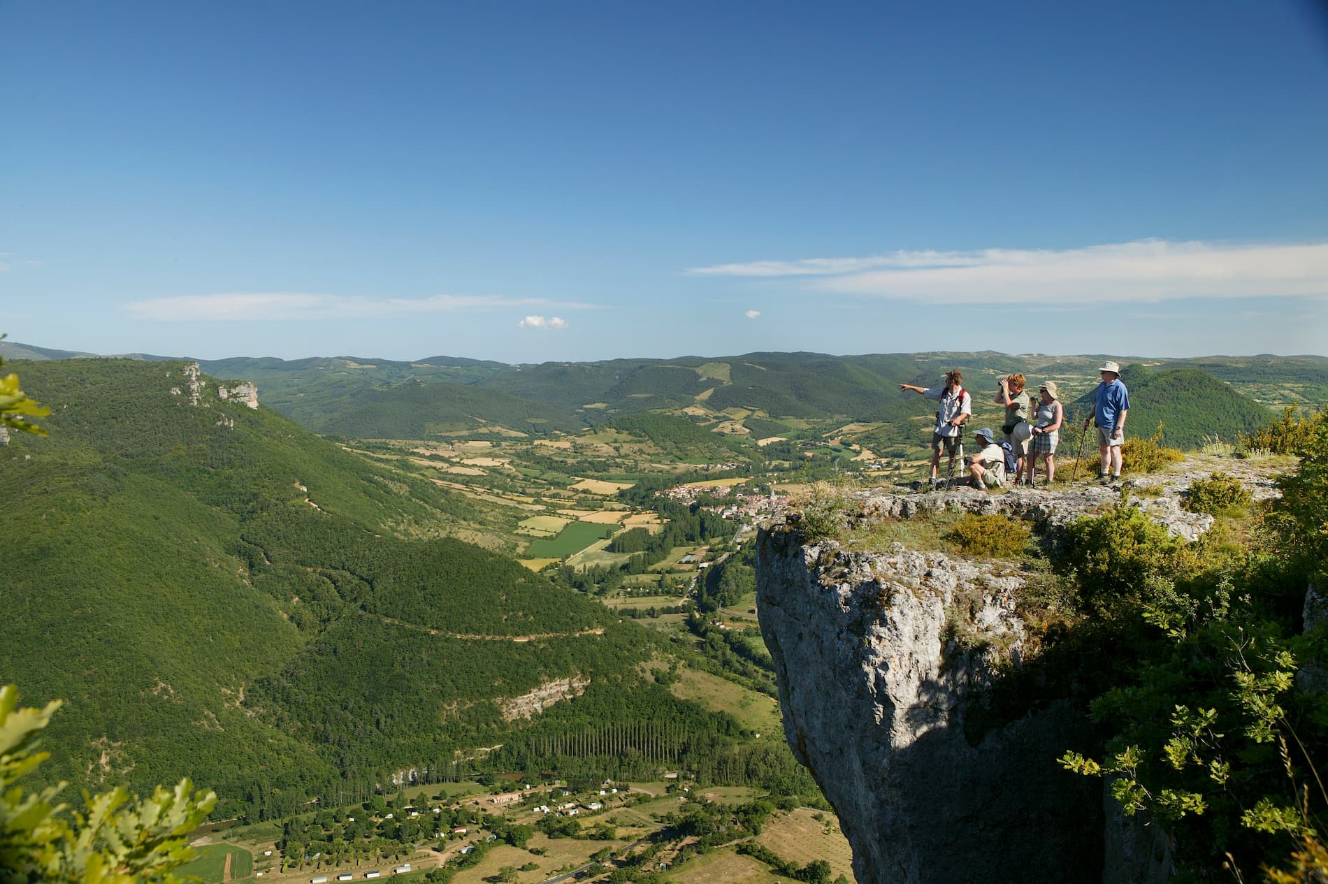 Randonneurs debout au bord d’une falaise rocheuse surplombant une vallée verdoyante et un village, sous un ciel bleu.