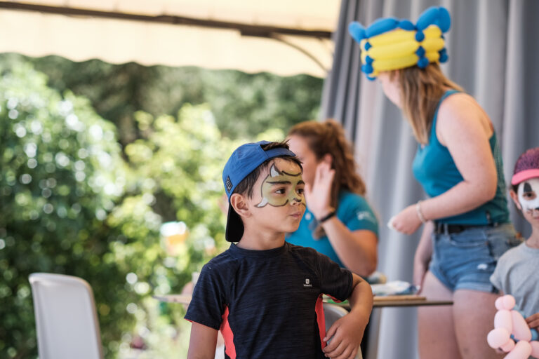 Enfant souriant avec maquillage de personnage (masque et moustache) portant une casquette bleue, dans un atelier extérieur avec d’autres enfants déguisés.