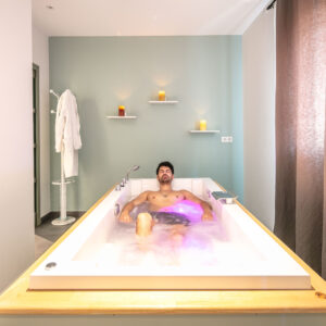 Homme nu allongé dans une baignoire en spa, éclairage violet, dans une salle de bain moderne avec étagères murales, rideau sombre et serviette sur un porte-linge.