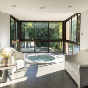 Espace spa intérieur lumineux avec deux fauteuils blancs, une petite table, une vasque à bulles, et une grande baie vitrée donnant sur la verdure.