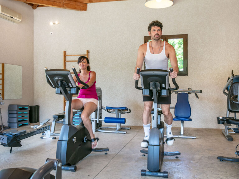 Deux personnes faisant du vélo d’intérieur dans une salle de sport, la femme à gauche et l’homme à droite sur des machines cardio.