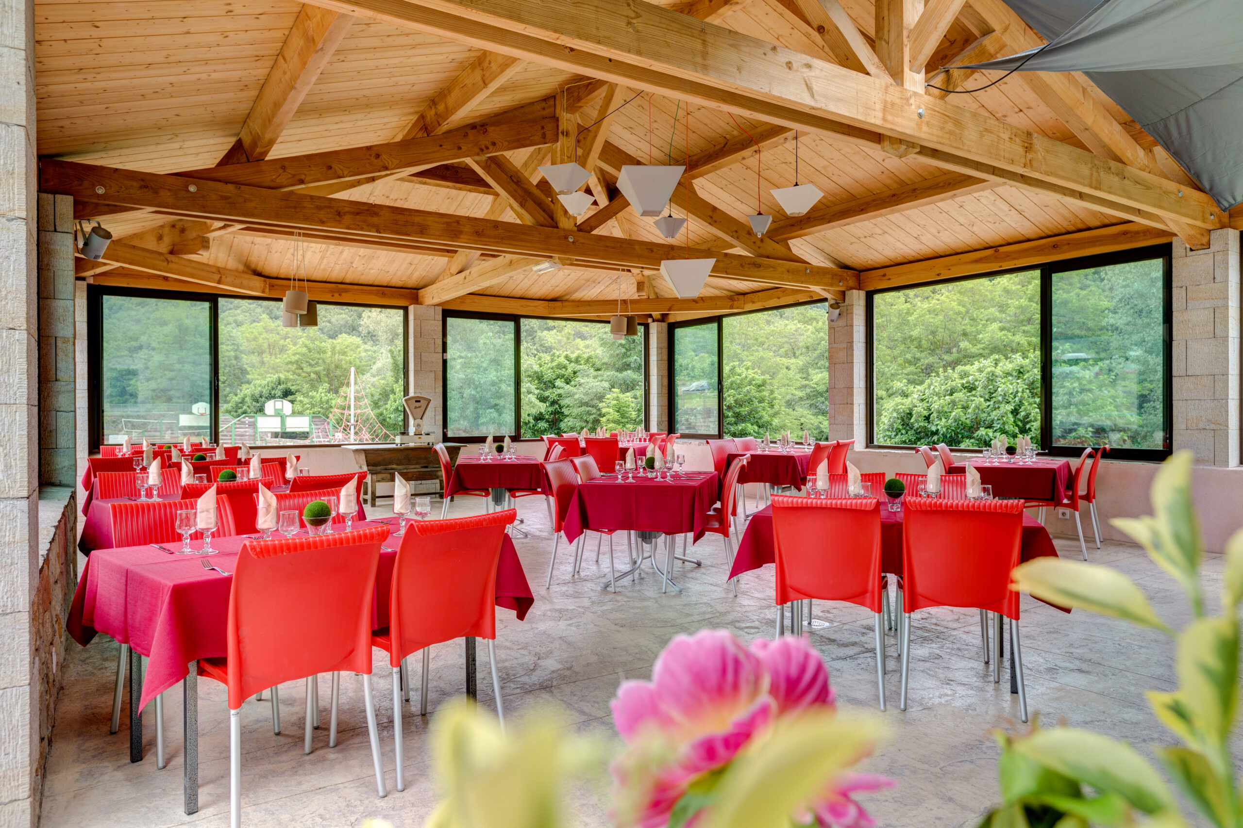 Salle de réception couverte avec tables dressées en rouge, chaises métalliques et décorations, fenêtres panoramiques donnant sur la verdure, poutres en bois au plafond et suspensions lumineuses.